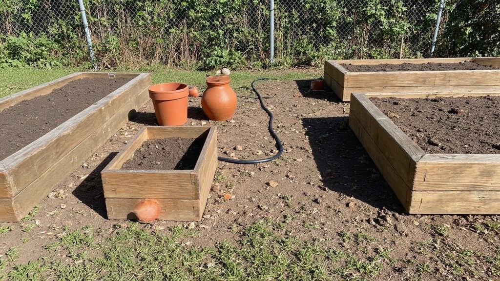 Overexposed afternoon photograph showing three rectangular raised garden beds constructed from weathered cedar planks, positioned on patchy grass with harsh shadows casting across uneven soil. Several terra cotta containers of varying sizes scattered nearby, some tipped at awkward angles. A basic drip irrigation system partially visible with black tubing snaking between beds. Background features a chain-link fence with climbing weeds, creating a utilitarian beginner gardening setup.